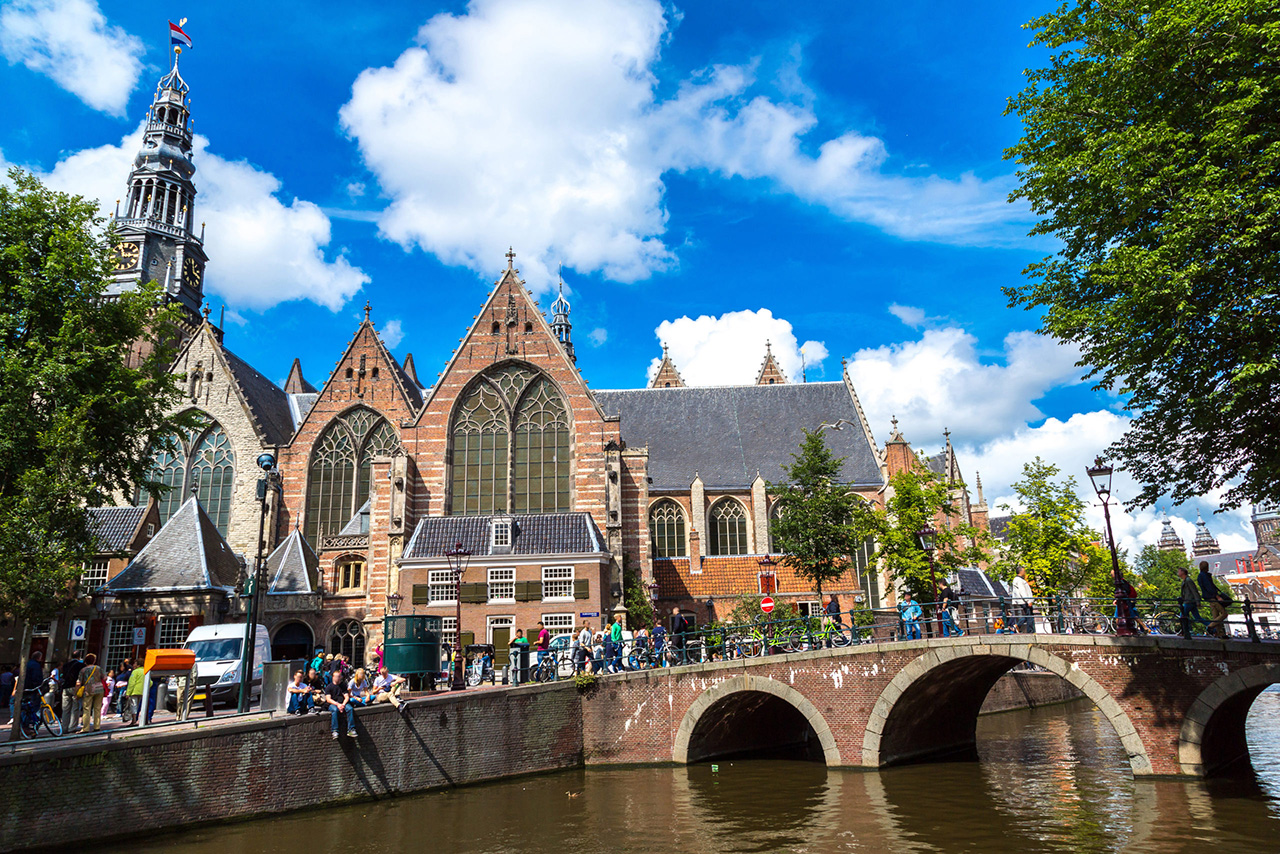 Iglesia Oude Kerk y su torre con bandera holandesa junto a un canal en Ámsterdam en un día soleado.
