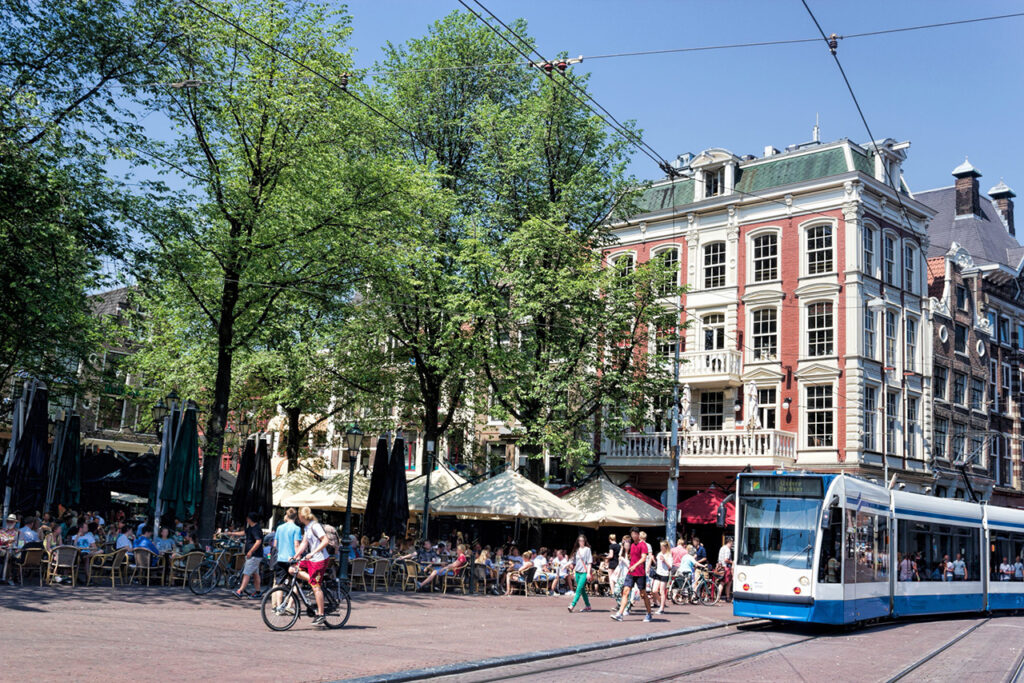 Tranvía y ciclistas en la animada plaza Leidseplein de Ámsterdam con terrazas de cafés y árboles verdes.